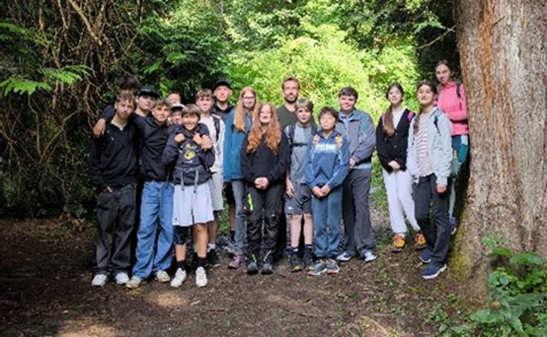 Eine Gruppe von Menschen posiert in einem Waldgebiet neben einem großen Baum. Sonnenlicht filtert durch das grüne Laub.