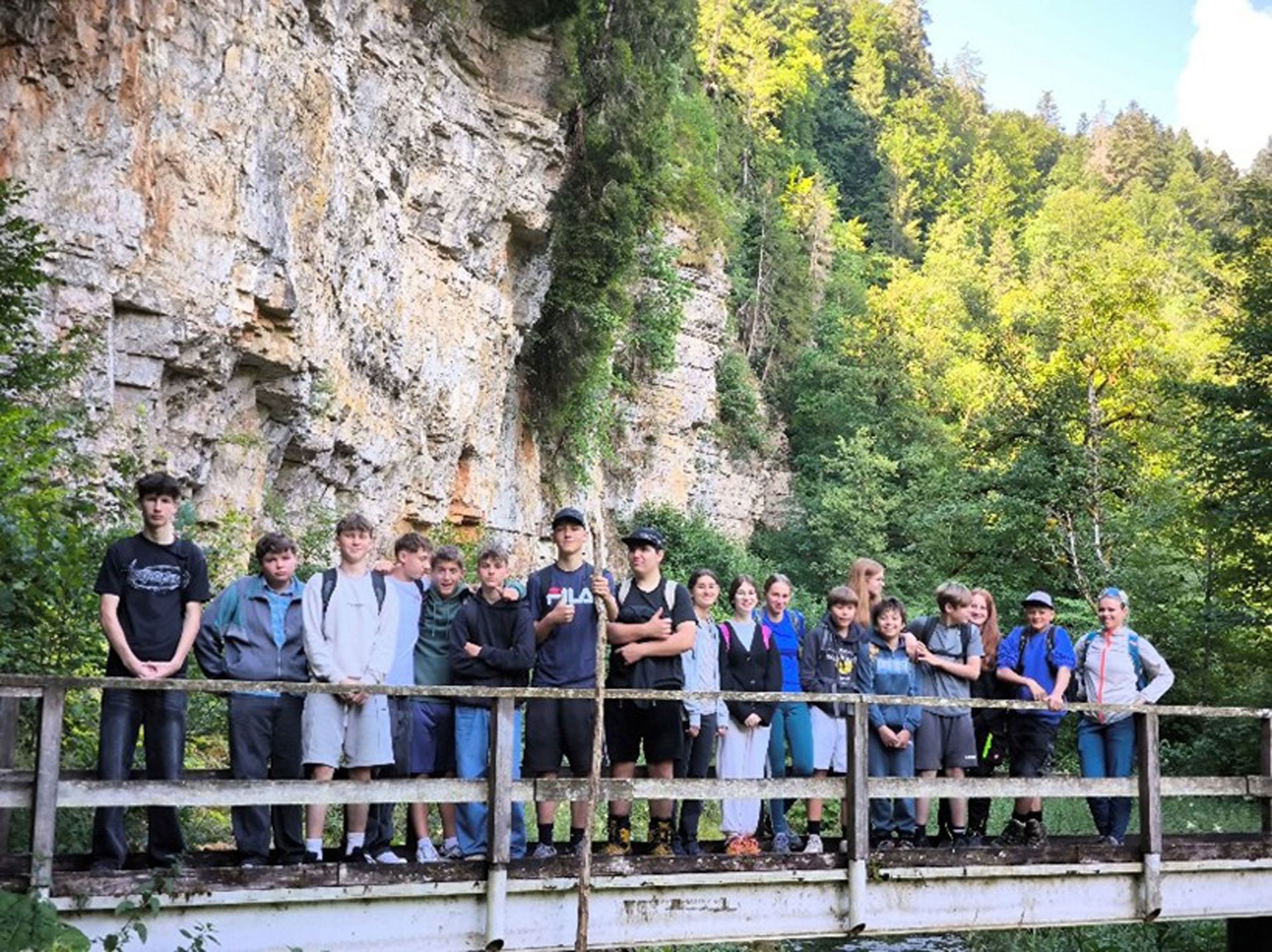 Eine Gruppe von Menschen steht auf einer Brücke vor einer Felswand und einem Wald.