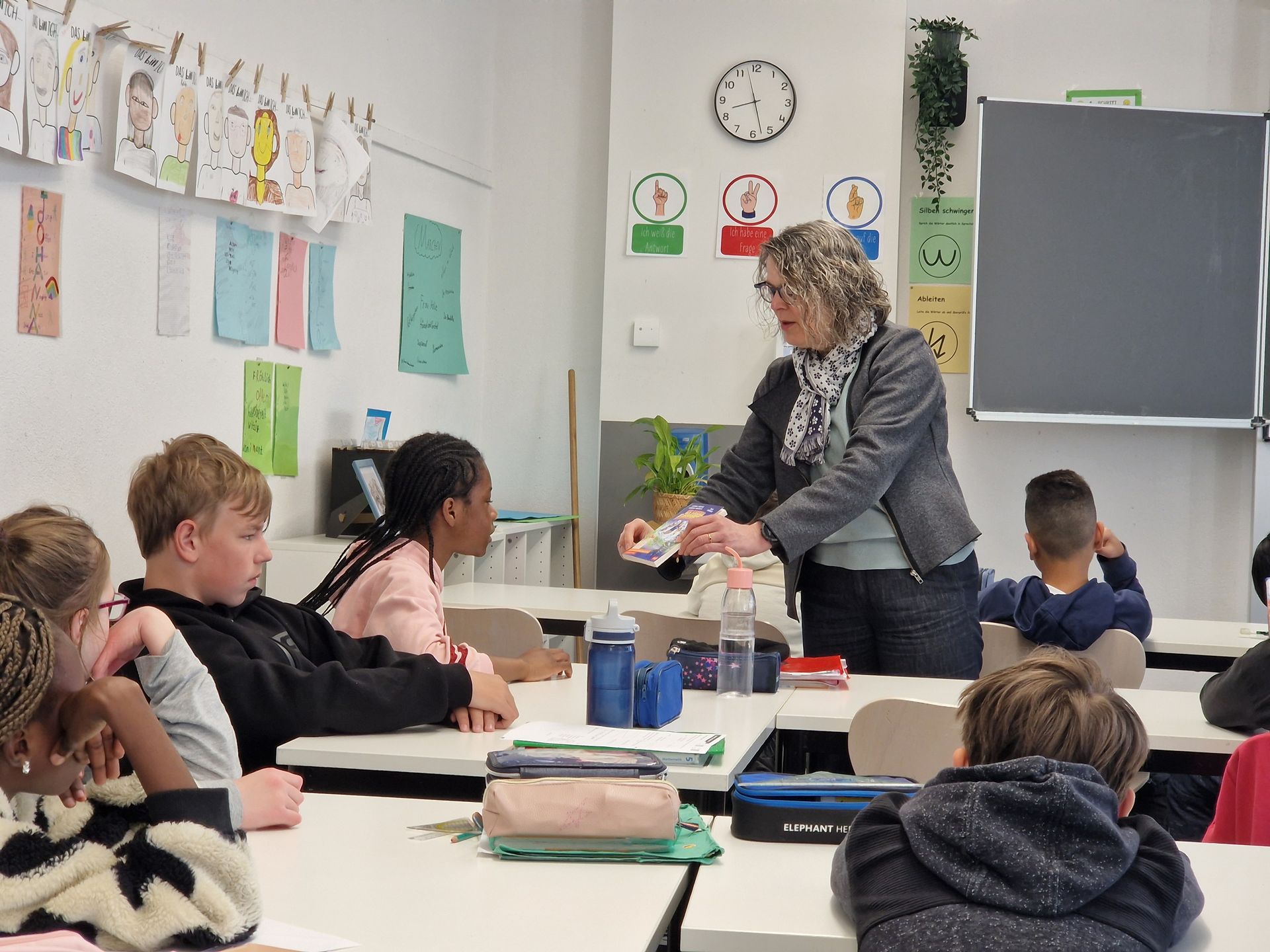 Teacher helping students at a classroom table, with clocks and papers on the wall.
