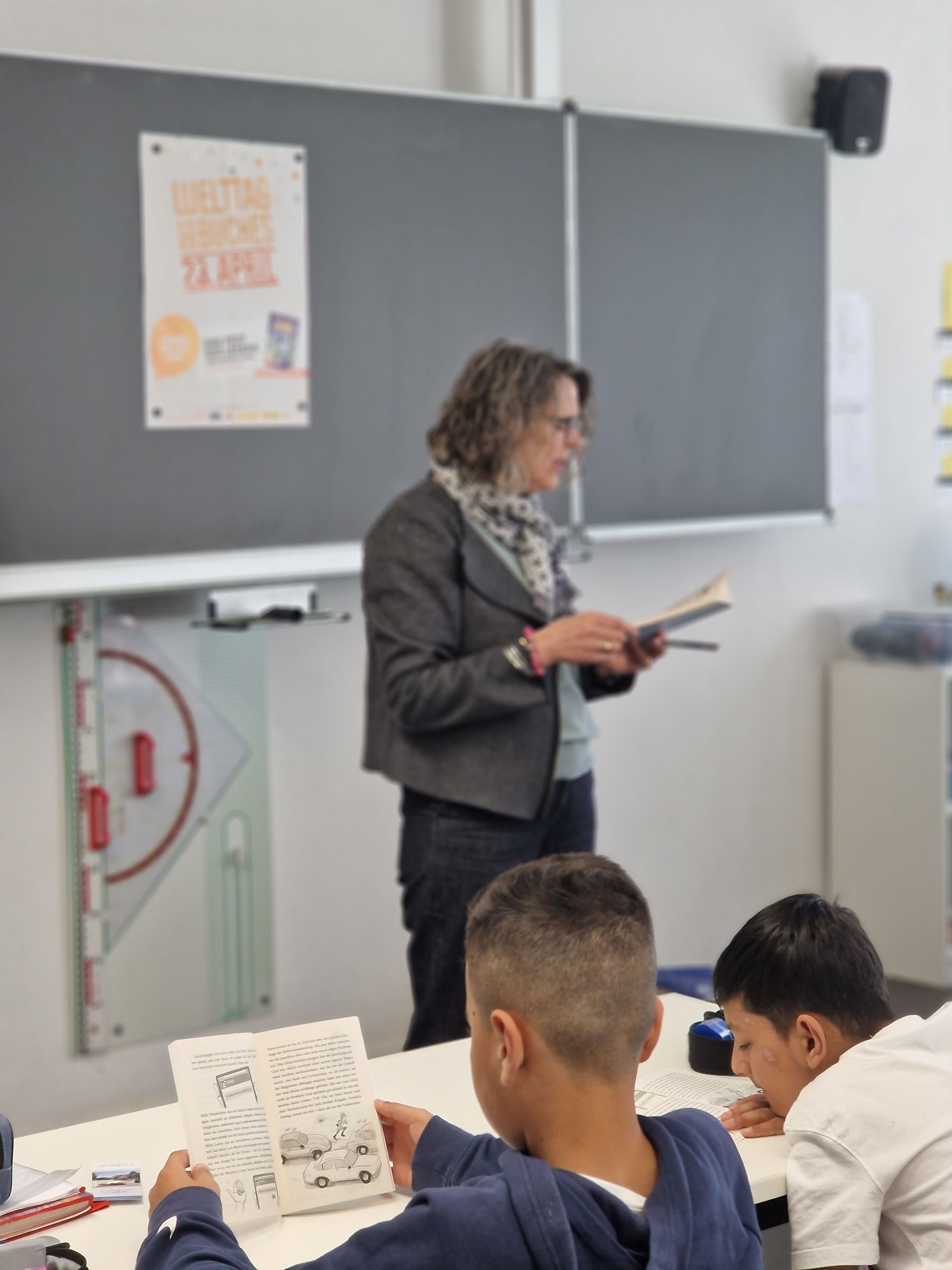 Teacher reading aloud in a classroom while students sit and read papers.