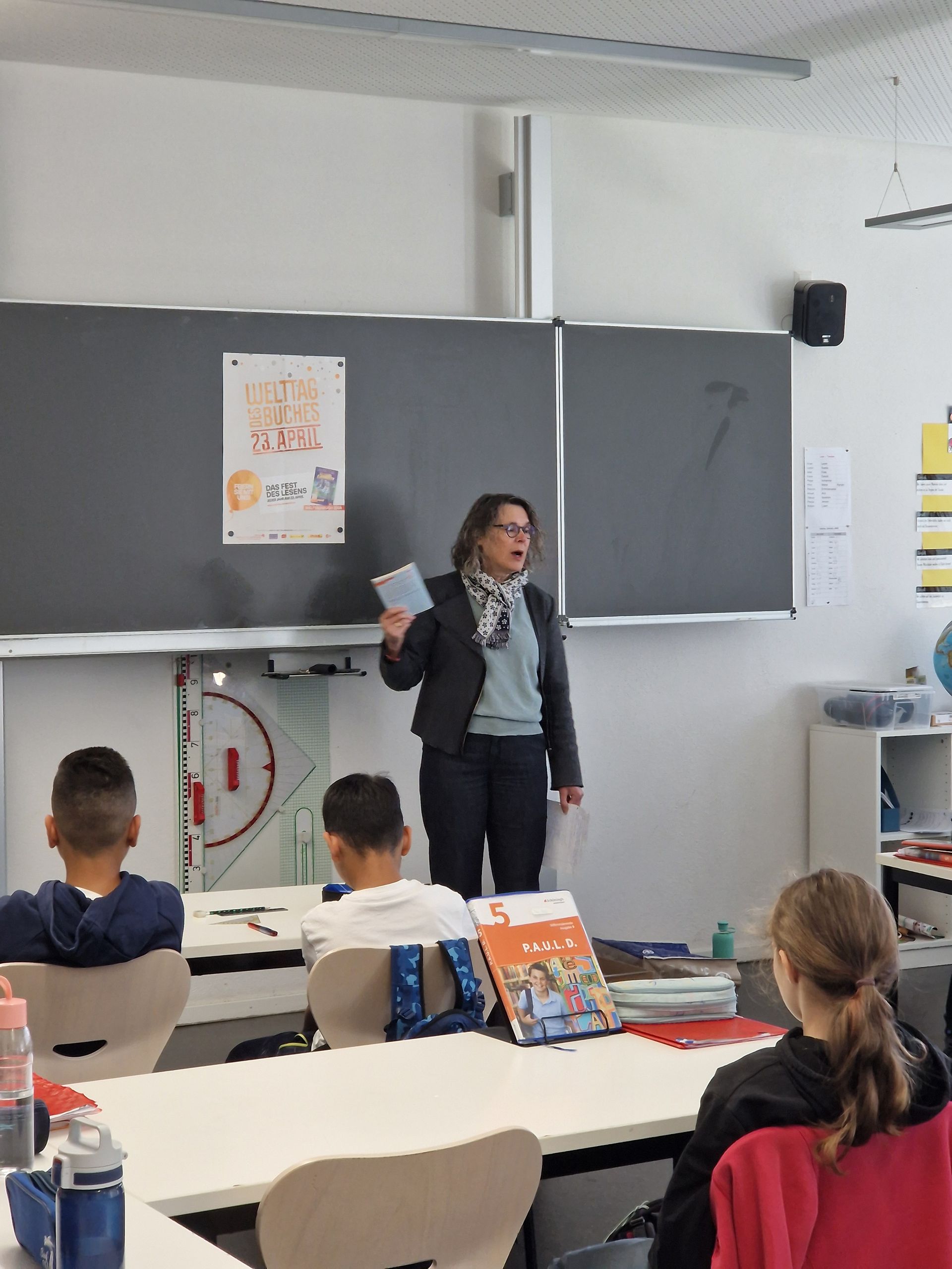 Teacher speaking at the front of a classroom while students sit at desks and watch.