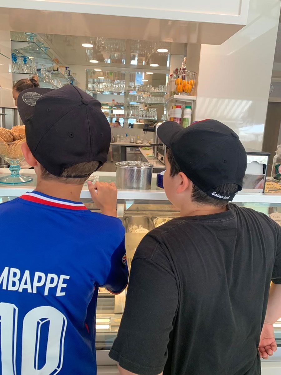 Zwei Personen mit schwarzen Kappen blicken in eine helle Auslage einer Bäckerei, eine von ihnen trägt ein blaues Mbappé-Trikot.