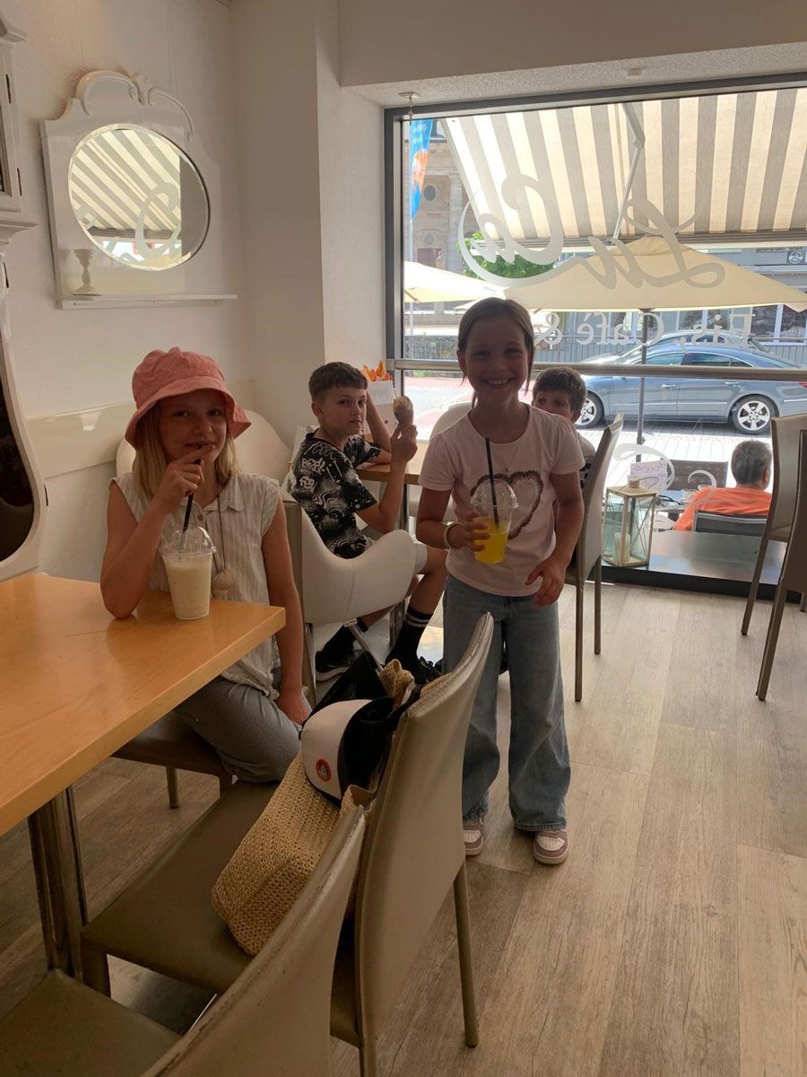 Kinder in einem hellen Café mit Getränken in der Hand, eines sitzt an einem Tisch, hinter ihnen ein großes Fenster.