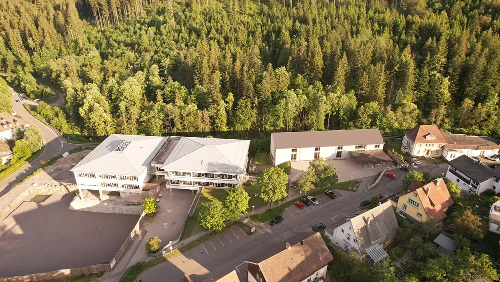 Luftaufnahme von Schulgebäuden neben einem Wald und einem Wohngebiet; sonniger Tag.