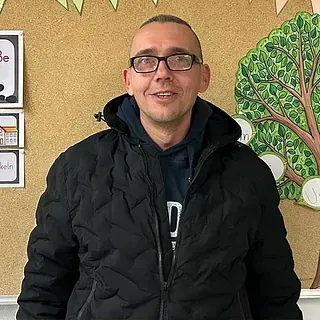 Ein Mann mit Brille und schwarzer Jacke lächelt in einem Klassenzimmer mit Pinnwand und Baumschmuck.