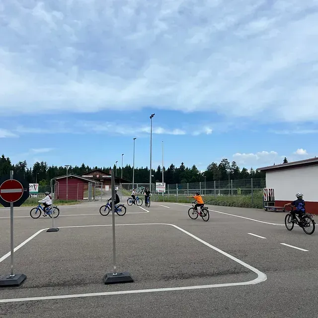 Kinder fahren auf Fahrrädern eine Strecke mit Verkehrsschildern ab, auf einem gepflasterten Platz mit Gebäuden, unter blauem Himmel.