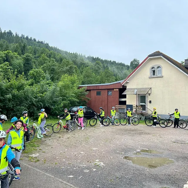 Eine Gruppe Kinder auf Fahrrädern, die Sicherheitswesten tragen, vor einem Gebäude und einem bewaldeten Hang.