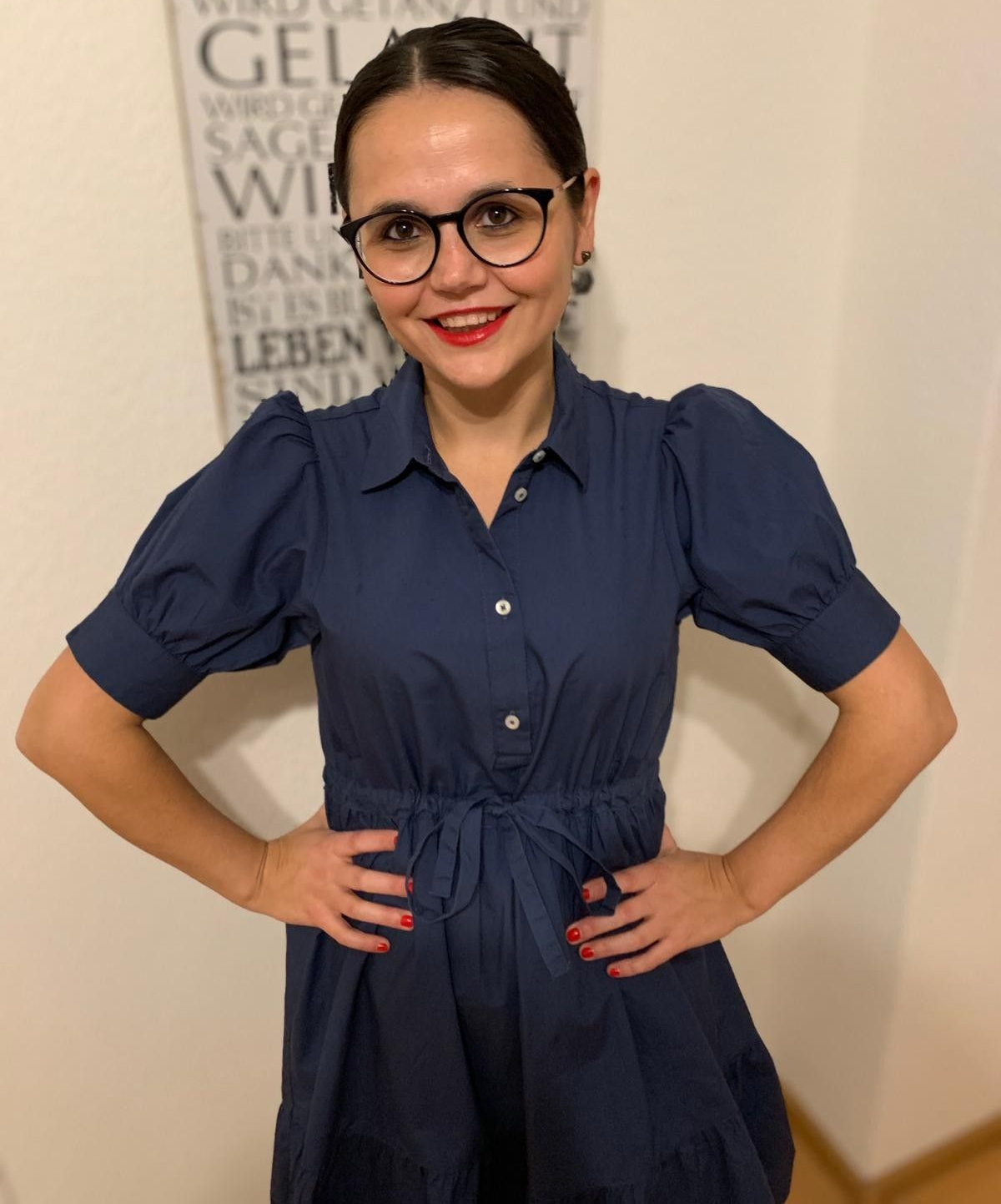 Frau in blauem Kleid mit Brille, Hände in die Hüften gestemmt, lächelnd, vor einem gerahmten Wandbild.