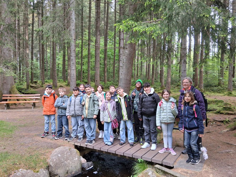 Group of children and adults posing on a wooden bridge in a forest clearing.