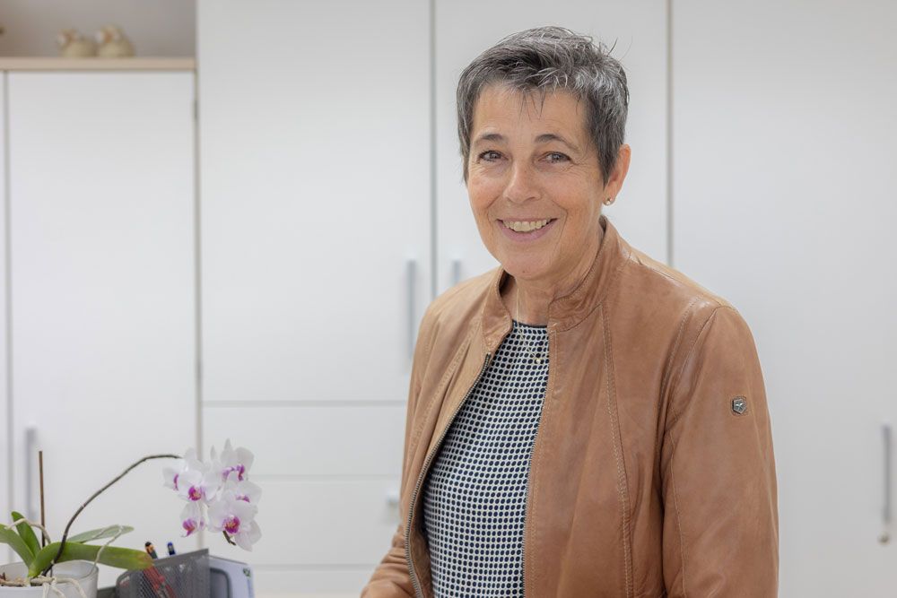 Eine Frau in brauner Jacke lächelt vor weißen Schränken; eine Orchidee ist zu sehen.