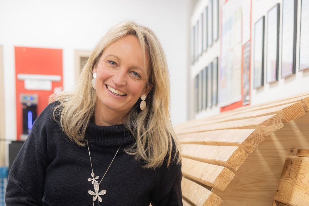 Eine lächelnde Frau lehnt an einer Holzbank. Sie trägt einen Pullover, eine Halskette und Ohrringe. Innenaufnahme bei hellem Licht.