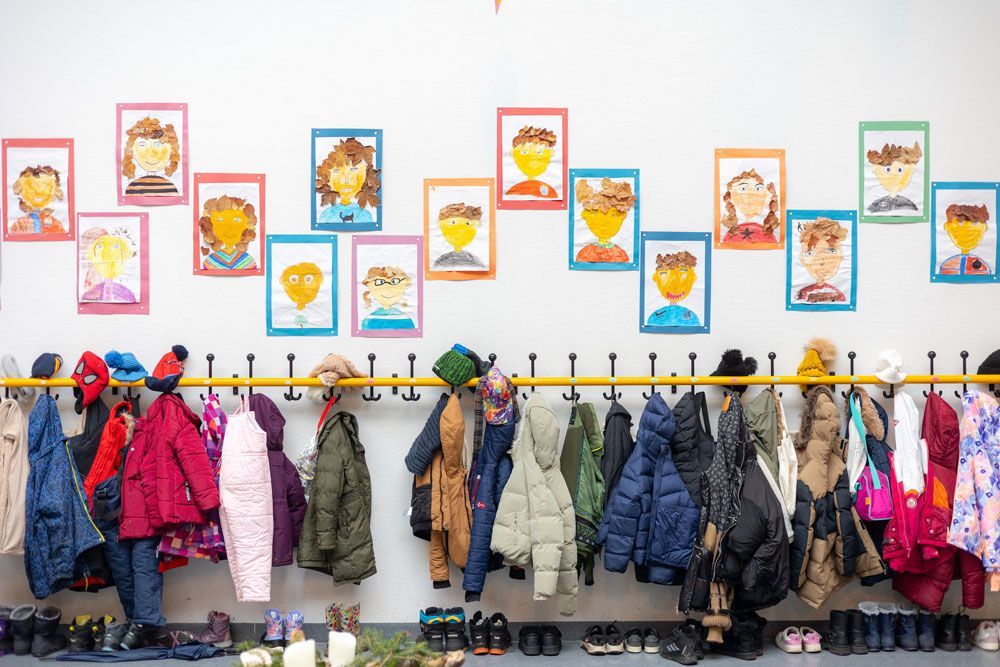 Eine Reihe bunter Kinderjacken und -schuhe hängt unterhalb von Kinderporträts an einer weißen Wand.