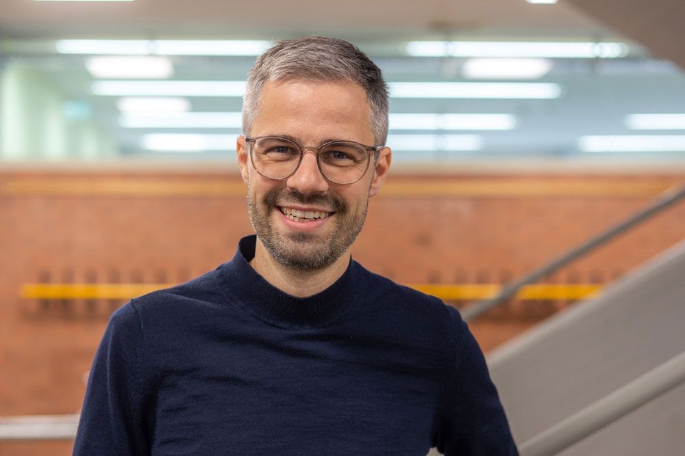 Ein Mann mit Brille und einem dunkelblauen Pullover lächelt und steht vor einer Backsteinmauer und einer Treppe.