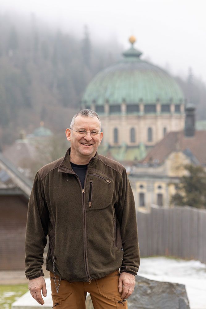 Ein lächelnder Mann in brauner Jacke und Hose steht an einem nebligen Tag vor einem Gebäude mit Kuppeldach.