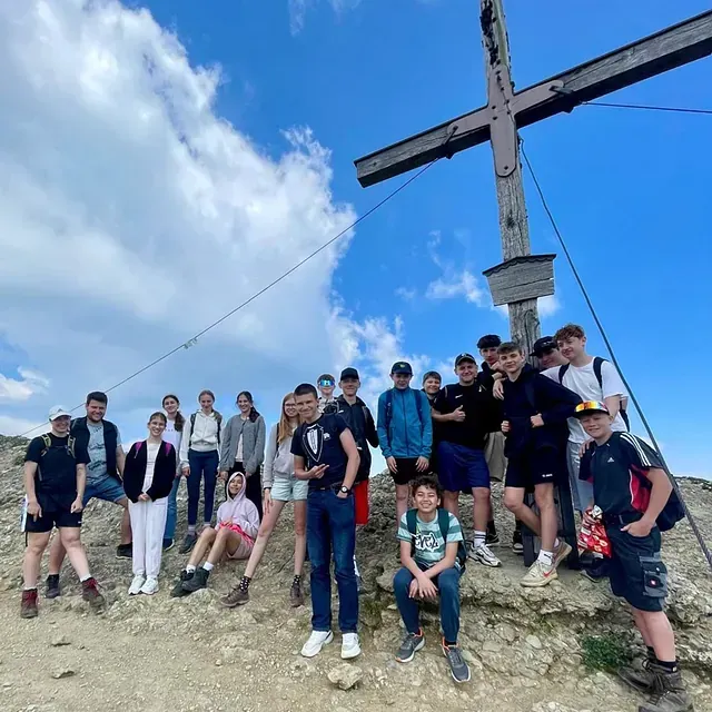Eine Gruppe von Menschen steht auf einem Berggipfel neben einem Holzkreuz. Im Hintergrund blauer Himmel mit Wolken.