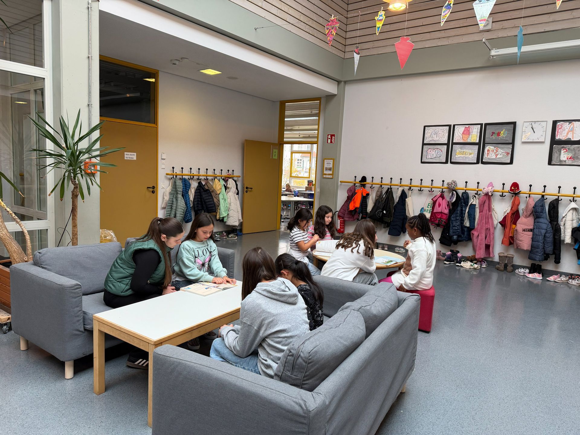 Kinder sitzen auf Sofas in einem Schulflur und spielen ein Spiel, Mäntel hängen an der Wand.