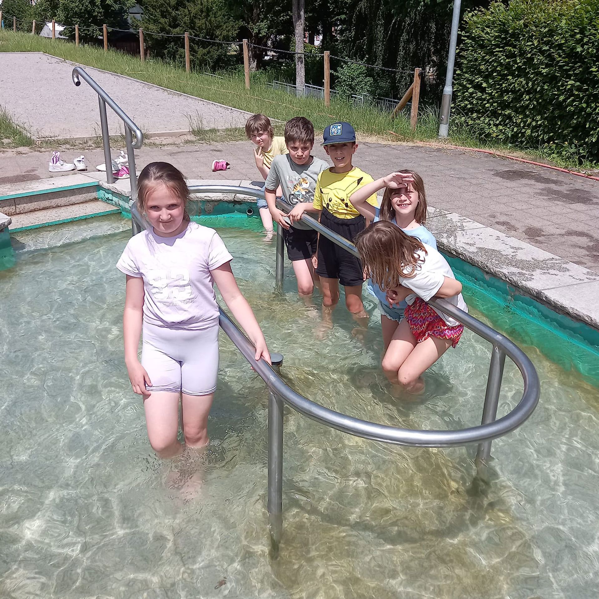 Kinder waten in einem flachen Becken und halten sich an einem Geländer fest. Sonniger Tag.