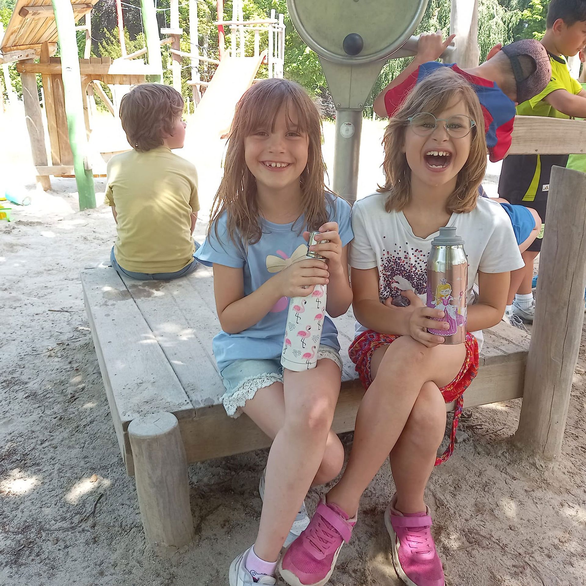 Zwei lächelnde Mädchen sitzen lachend auf einer Holzbank, halten Wasserflaschen in den Händen. Spielplatzszene.