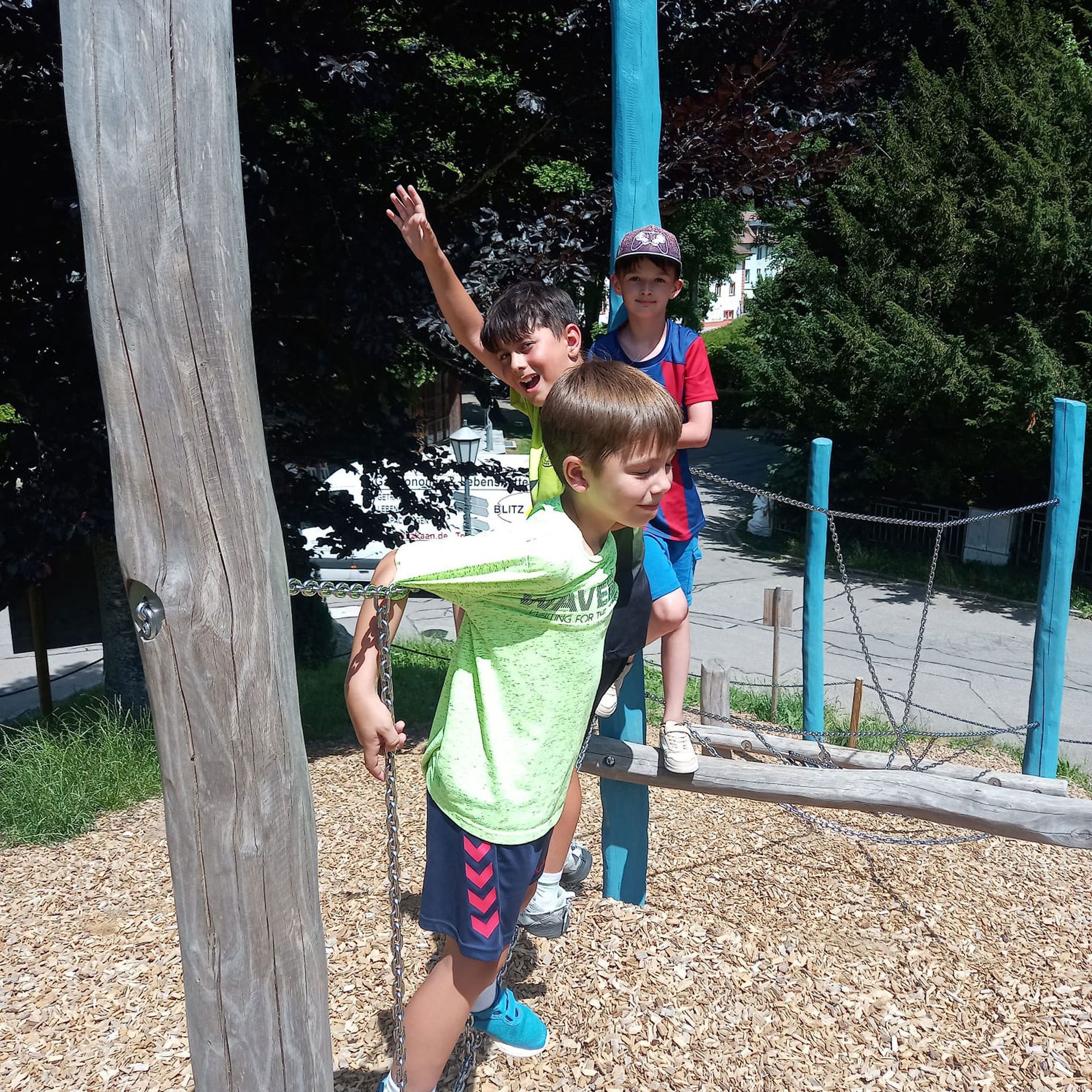 Drei Jungen spielen auf einem Spielplatz, einer hebt die Hand. Strahlend sonniger Tag.