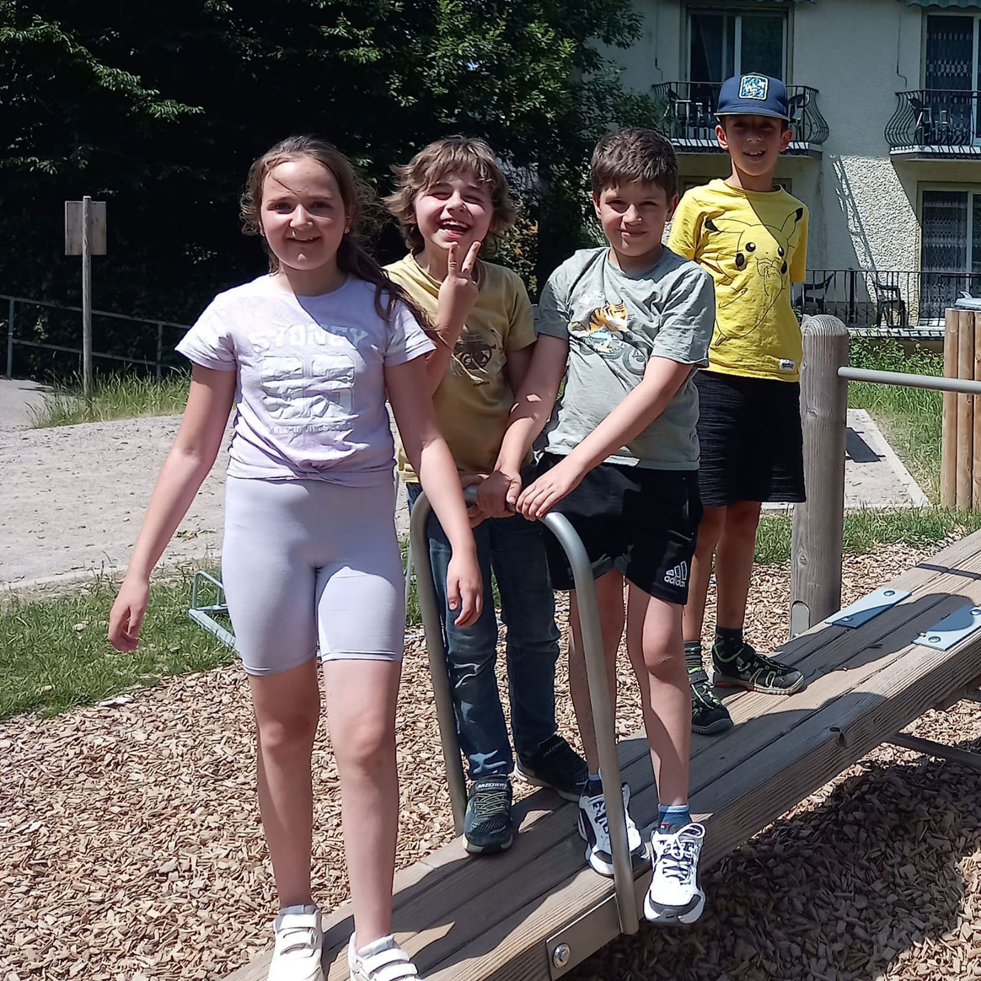 Vier Kinder auf einer Holzwippe auf einem Spielplatz, posieren und lächeln an einem sonnigen Tag.
