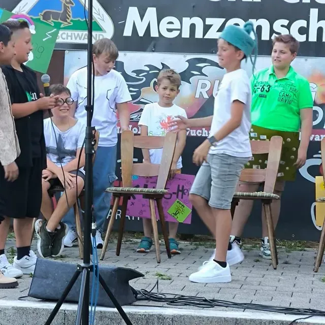 Kinder spielen Stuhltanz auf einer Bühne; einige stehen, andere sitzen. Zwei Stühle sind zu sehen.