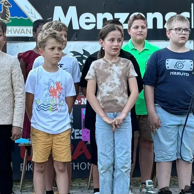Eine Gruppe Kinder steht vor einem Schild. Ein Mädchen in einem Batik-Shirt und Jeans schaut geradeaus. Die anderen schauen in die Kamera.