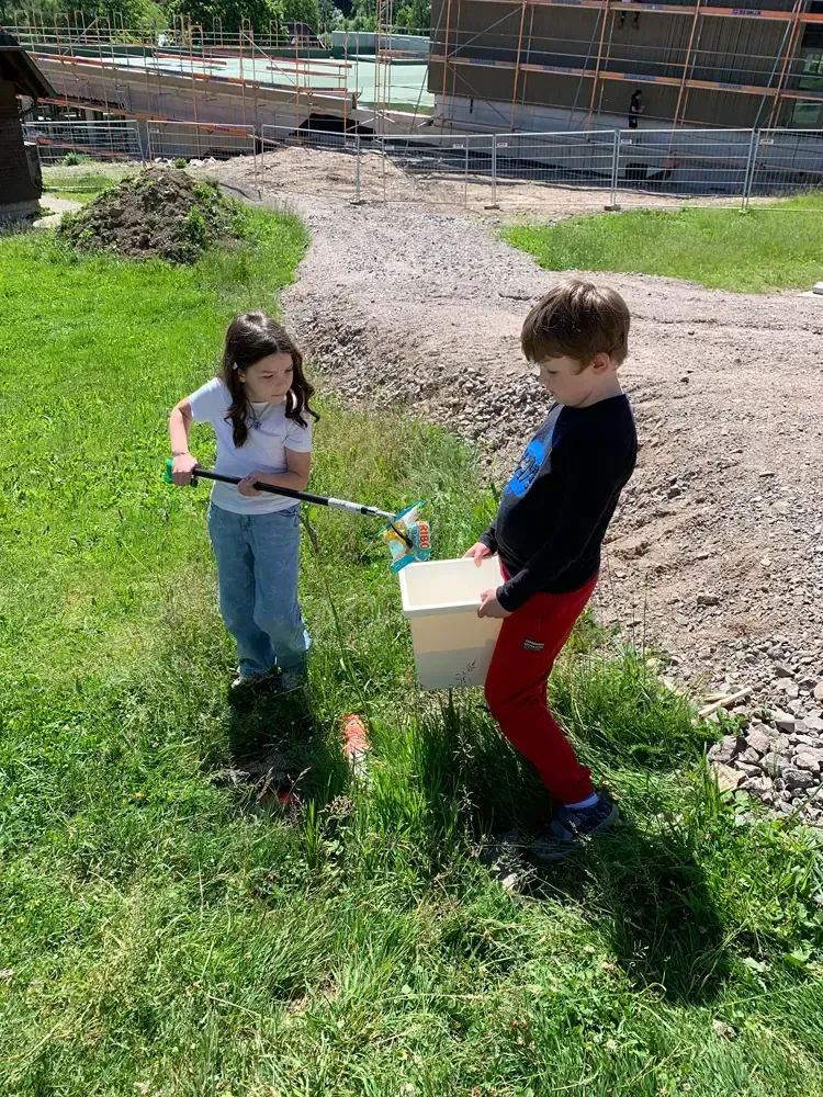 Two children playing with colorful hose sprayers on a grassy slope near a construction site.