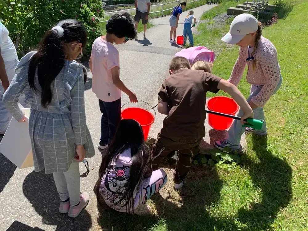Children gather around red buckets on a sunny path, scooping water near a grassy area.