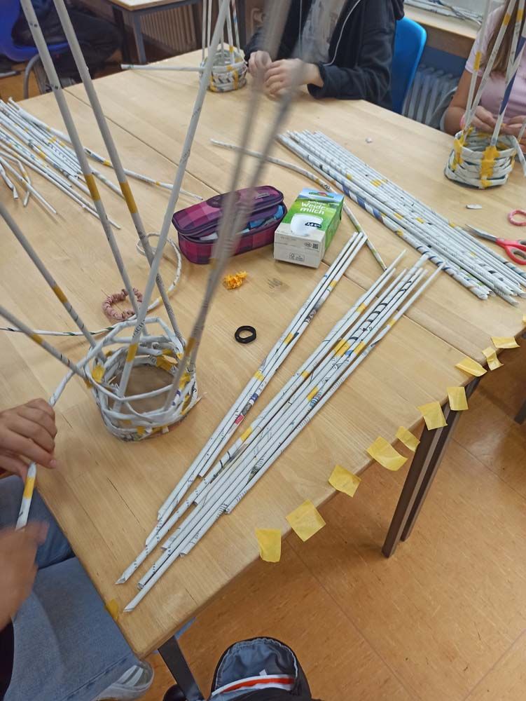 An einem Tisch flechten Menschen Körbe aus Papierröhren. Material, Scheren und ein Saftkarton sind ebenfalls vorhanden.