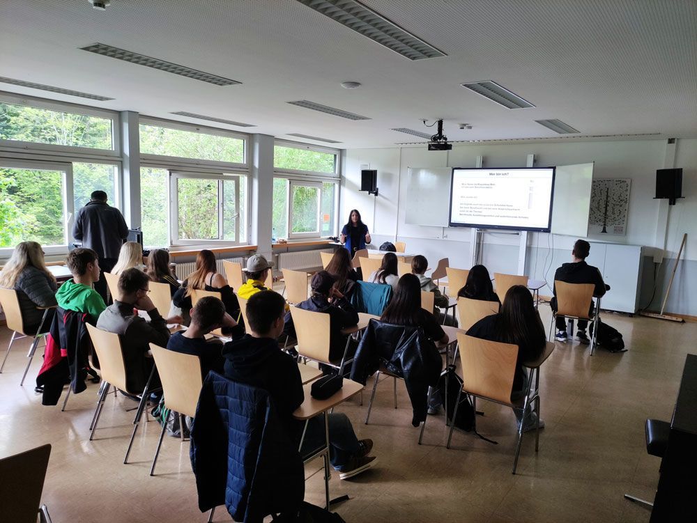 Präsentation im Klassenzimmer mit sitzenden Zuhörern, die in einem hellen Raum auf eine Leinwand schauen.