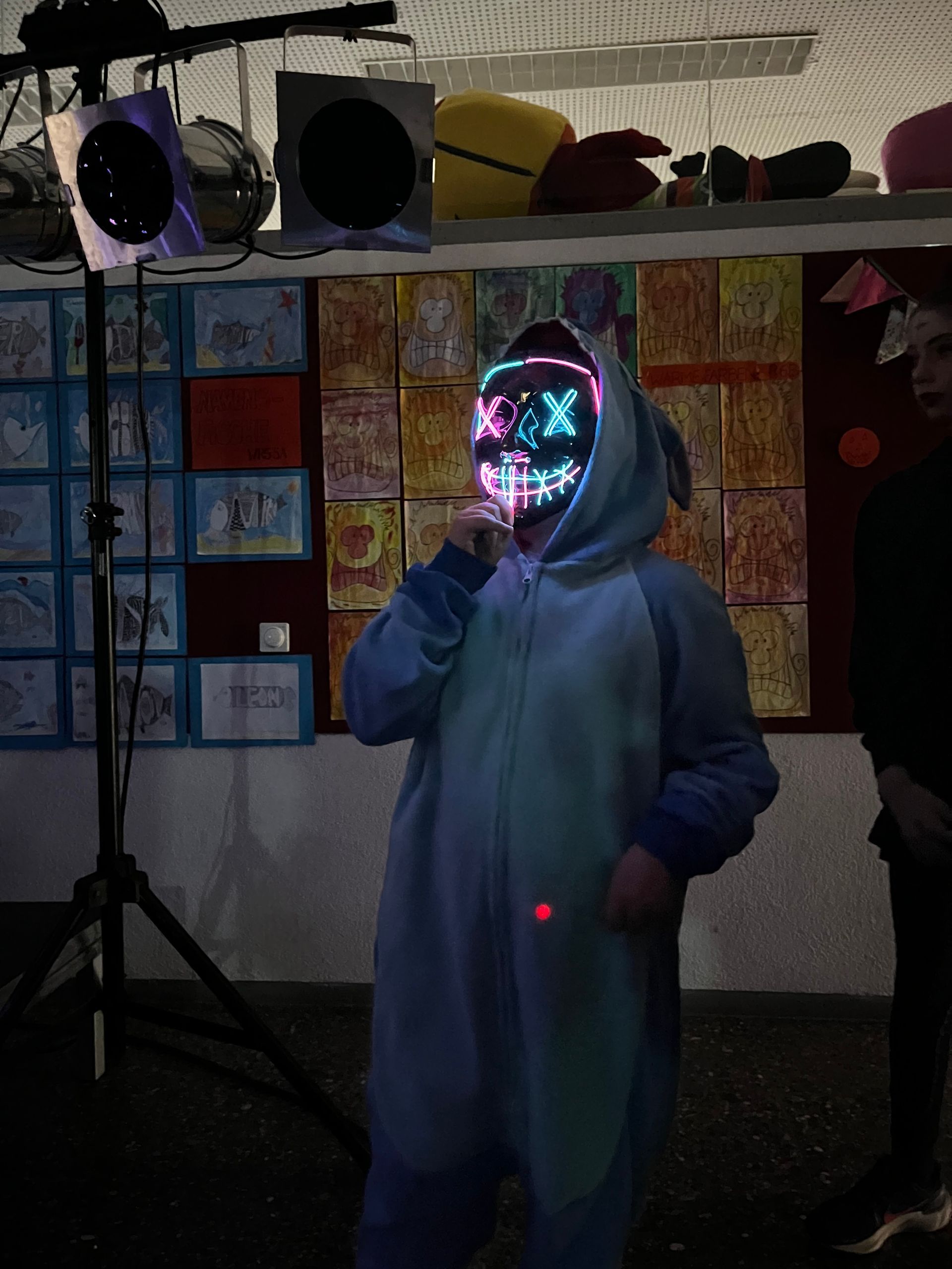 Eine Person im Stitch-Einteiler mit leuchtender Maske steht in der Nähe einer Bühne; im Hintergrund ist ein Kunstwerk zu sehen.