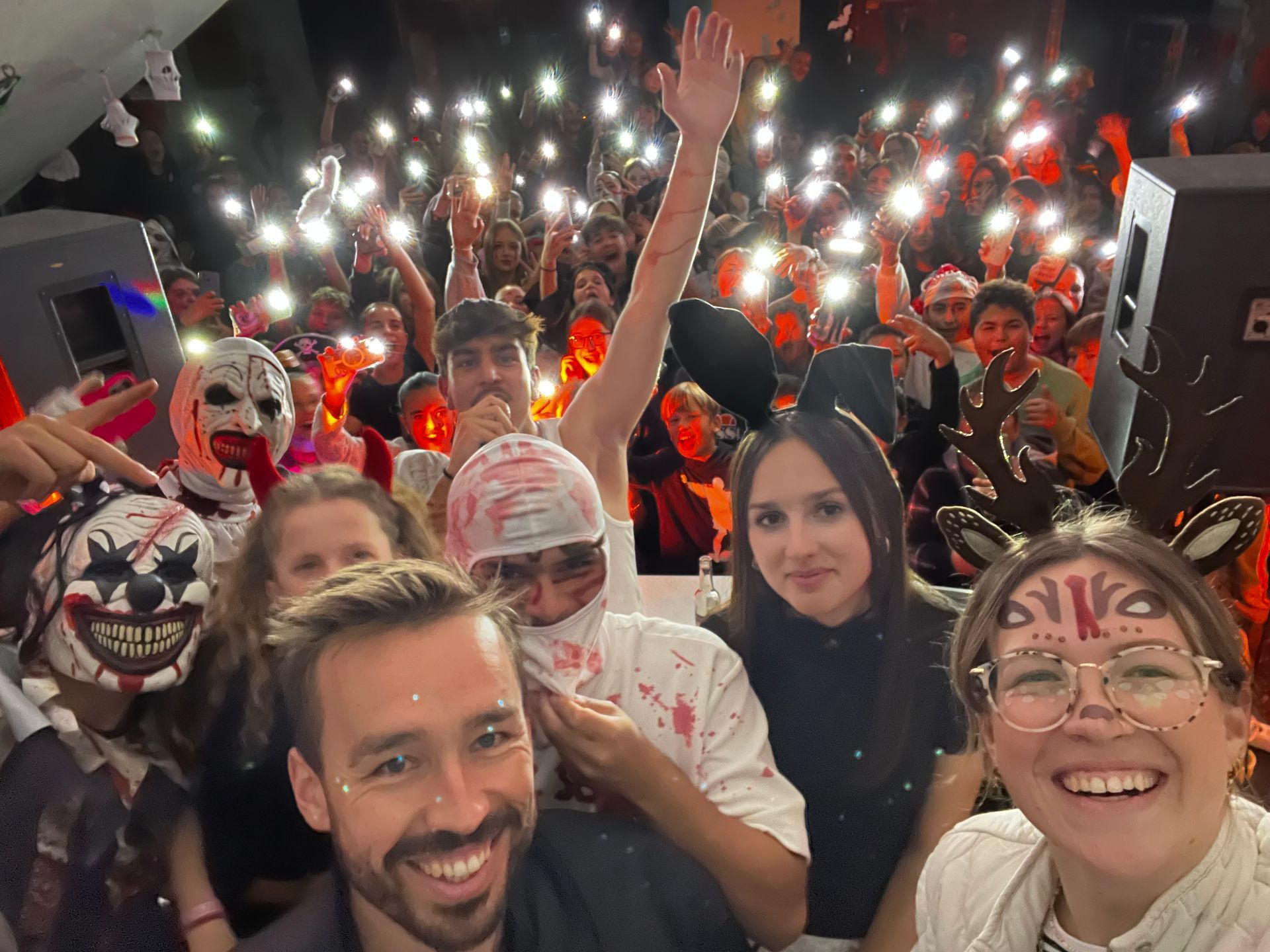 Menschen auf einer Party, einige in Kostümen, halten ihre Handys mit leuchtenden Lichtern hoch. Im Hintergrund ist ein DJ-Pult zu sehen.