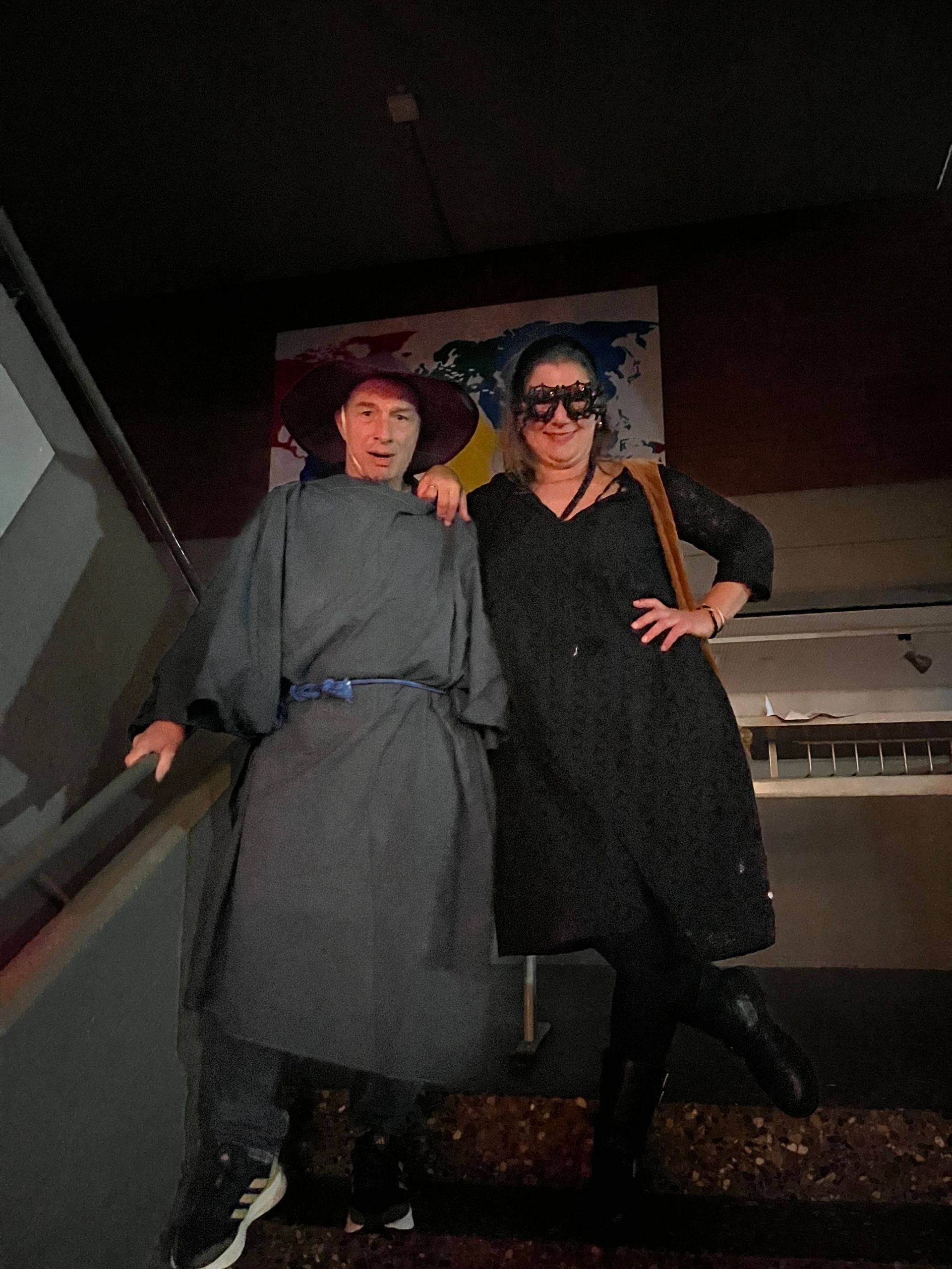 Zwei Personen auf einer Treppe; eine in grauer Kleidung mit Hut, die andere in schwarzem Kleid und Maske. Im Hintergrund ein Wandgemälde.