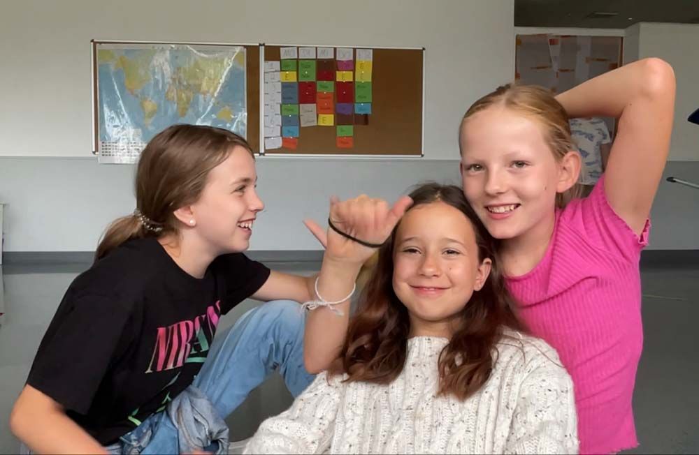 Drei lächelnde Mädchen posieren in einem Klassenzimmer; eines zeigt mit dem Daumen nach oben.