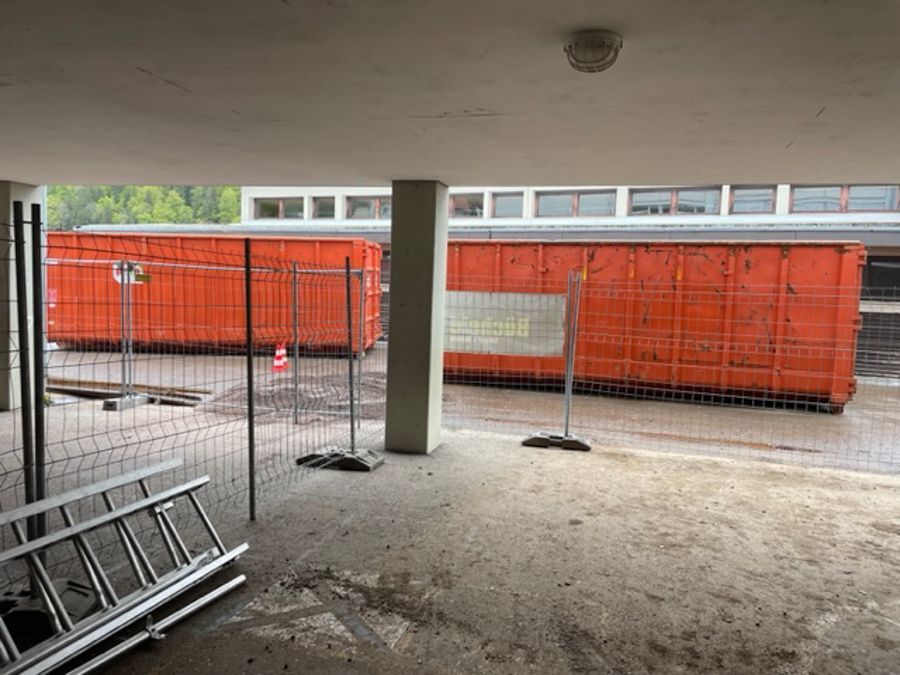 Zwei orangefarbene Abfallcontainer hinter einem Zaun, unter dem Dachvorsprung eines Gebäudes.