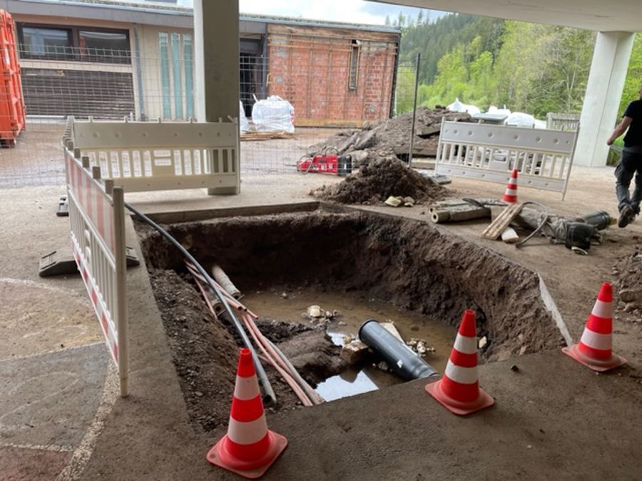Baustelle mit einer tiefen Baugrube, Sicherheitskegeln und freiliegenden Rohren unter einem Gebäude.