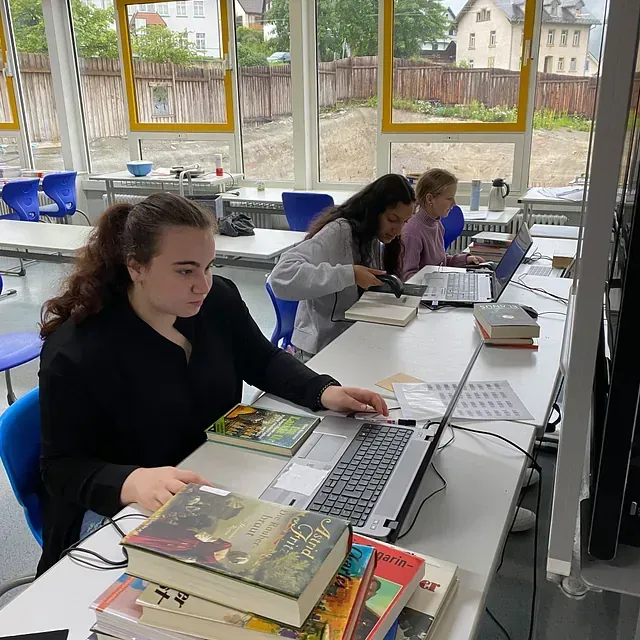 Drei Personen arbeiten an einem langen Tisch in einem Raum mit großen Fenstern; Bücher und Laptops sind vorhanden.