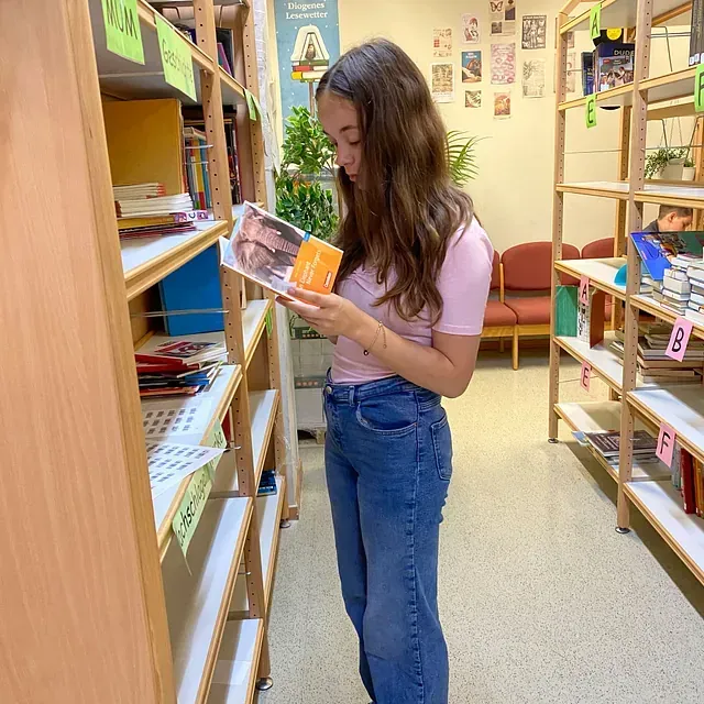 Eine Frau in rosa Hemd und Jeans betrachtet ein Buch in einer Bibliothek. Holzregale und Bücher umgeben sie.