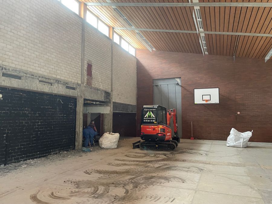 Bauarbeiten in der Turnhalle mit Bagger, Ziegelwänden, Basketballkorb und zwei Arbeitern.