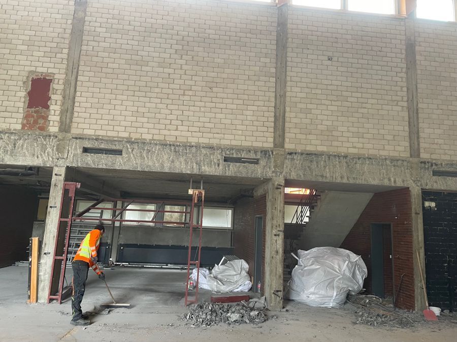 Ein Bauarbeiter fegt den Boden in einem Gebäude, das renoviert wird. Betonpfeiler stützen die Ziegelwände.