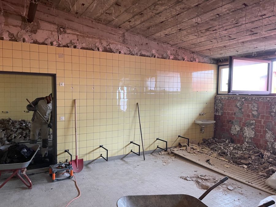 Raum im Abrisszustand; gelbe Fliesenwand, Schutt, Werkzeuge, Arbeiter sichtbar.