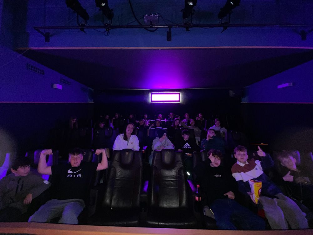 Dark theater audience seated in rows under purple lighting, watching a screen.