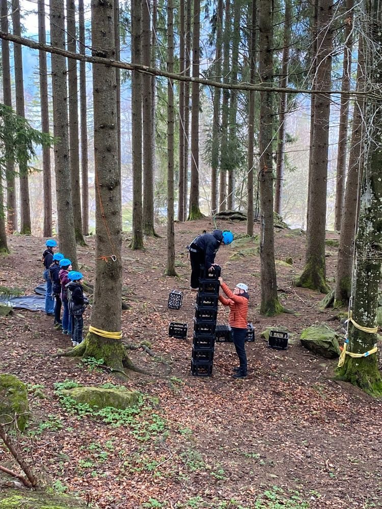 People in helmets stack black blocks in a pine forest setting