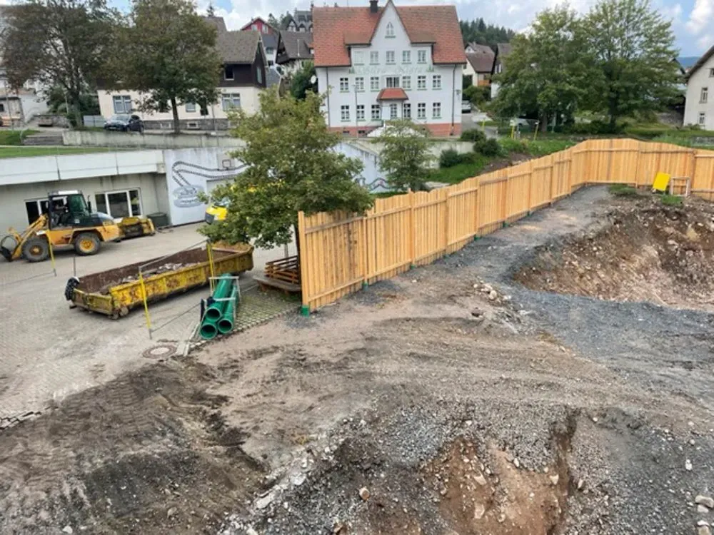Baustelle mit Holzzaun, gelbem Traktor und Gebäuden im Hintergrund.