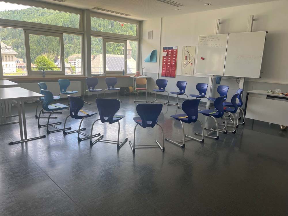 Ein Klassenzimmer mit blauen Stühlen, die im Kreis vor einer Tafel angeordnet sind; die Fenster bieten Blick auf eine grüne Landschaft.