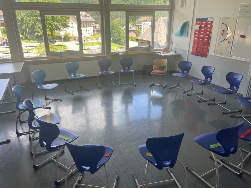Stühle sind in einem Klassenzimmer im Kreis angeordnet, draußen vor dem Fenster hat man Blick auf eine Stadt.