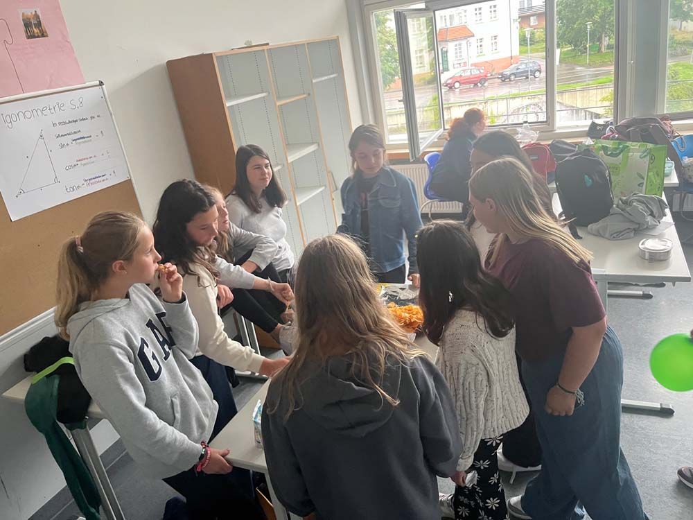 Eine Gruppe Mädchen sitzt in einem Klassenzimmer um einen Tisch und unterhält sich. Ein grüner Ballon und eine Tafel sind zu sehen.