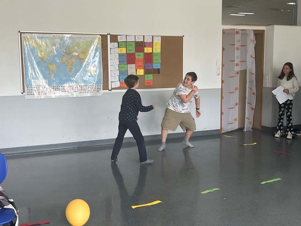 Zwei Jungen spielen Theater. Der eine schwingt den Arm, der andere reagiert. Ein Mädchen beobachtet sie mit Papieren in der Hand. An der Wand hängt eine Weltkarte.