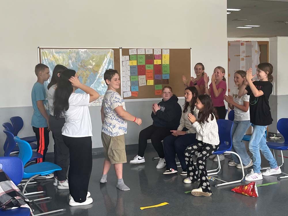 Schüler in einem Klassenzimmer klatschen und jubeln einem Schüler vor einer Weltkarte und einer bunten Tafel zu.