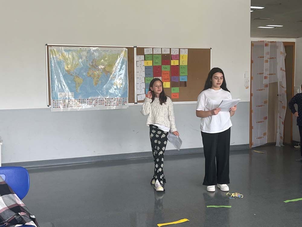 Zwei Mädchen präsentieren in einem Klassenzimmer vor einer Weltkarte und einer farbcodierten Tafel.